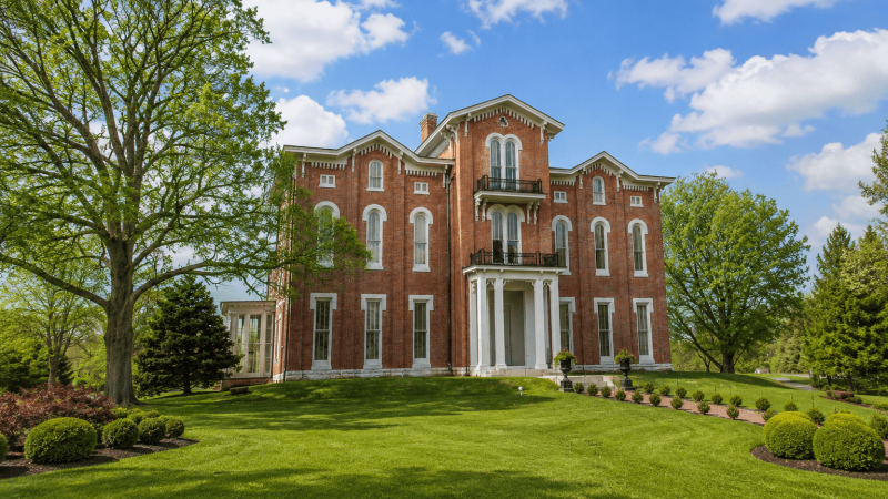Historic red-brick house with large trees, under a blue sky with clouds.