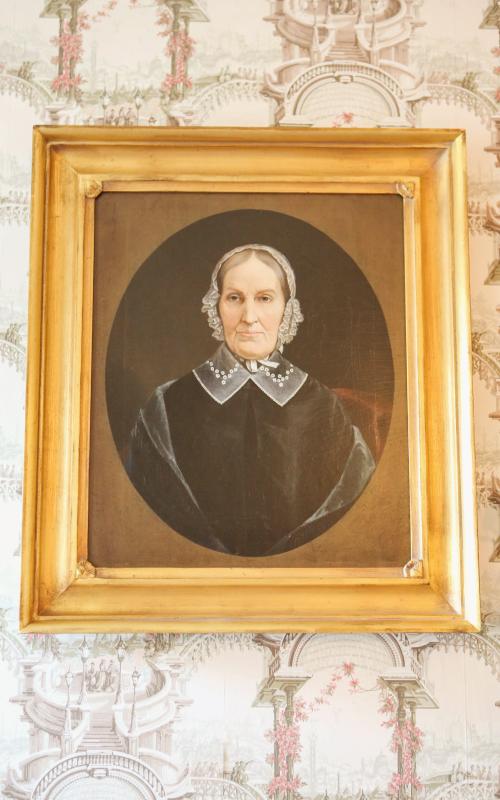 Framed portrait painting of an older person on ornate wallpaper background.
